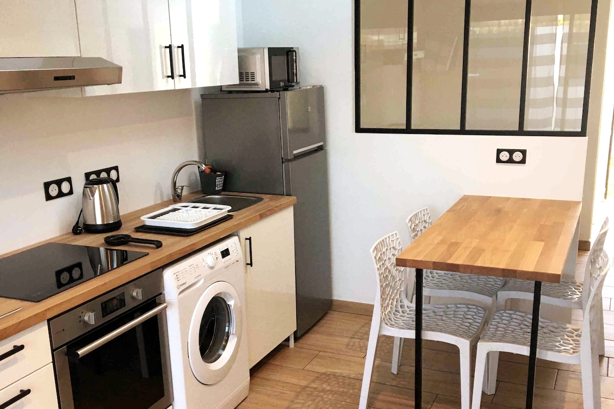 The kitchen area features modern appliances including a fridge, induction cooktop, and washing machine. A small table with four white chairs is positioned near a window, allowing natural light into the space. Sleek cabinets and a stainless-steel microwave complete the functional design.