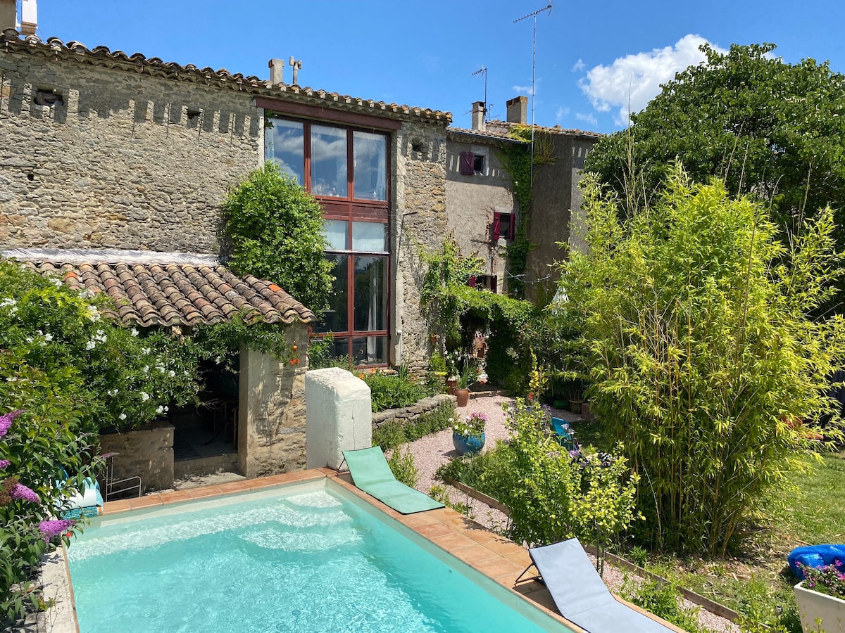A tranquil outdoor area is showcased, featuring a clear swimming pool surrounded by lush greenery. The stone building, with large windows, exudes a rustic charm. Sunlight casts a warm glow over the garden, creating an inviting atmosphere for relaxation.