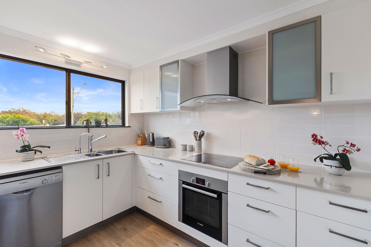 The kitchen features modern Bosch appliances, including a dishwasher, oven, and cooktop, complemented by ample counter space. Natural light enters through a large window, illuminating white cabinetry and decorative potted orchids. A neutral color palette promotes a functional and inviting cooking environment.