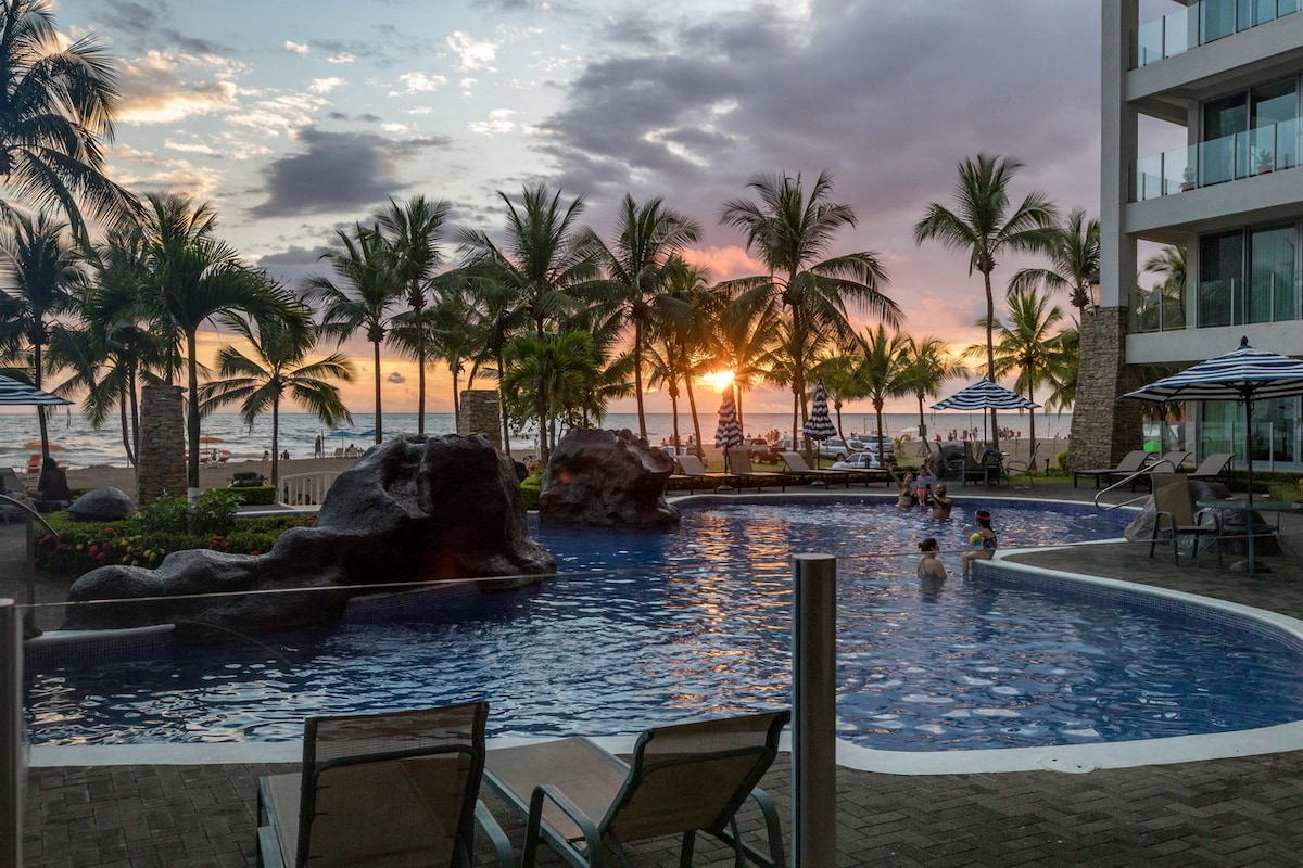A scenic view features a vibrant sunset over the ocean, with palm trees lining the shore. The inviting pool area includes natural rock formations and lounge chairs, creating a relaxed atmosphere. Guests can be seen enjoying the pool amid a tranquil backdrop of water and sky.