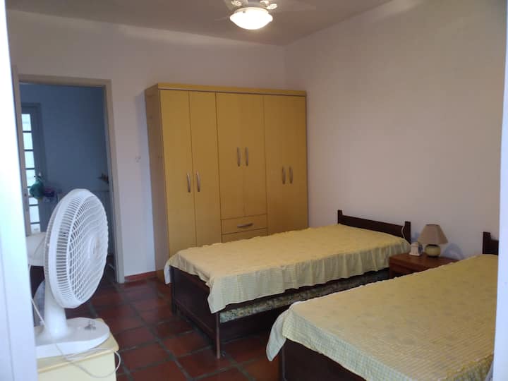 Dhomë gjumi në fund të shtëpisë me dollap, 2 krevate me trungje, ventilatorë tavani dhe tavoline.