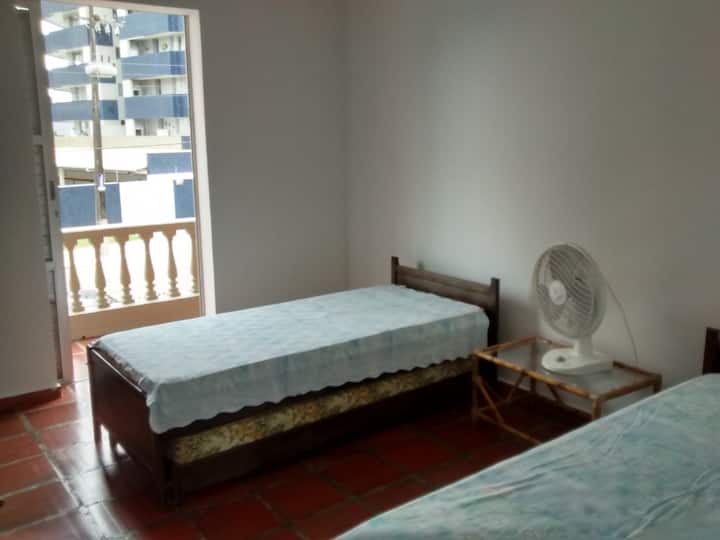 Dhomë gjumi e mesme me 2 krevate me trungje, me një dyshek në dysheme, dollap në mur që nuk shfaqet në fotografi dhe një ventilator tavoline.