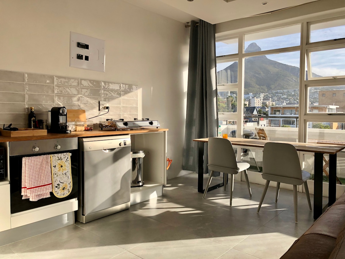 A bright kitchen space features modern appliances and a wooden countertop. Two light-colored chairs are positioned around a small table. A large window allows natural light to enter, framing a view of nearby mountains.