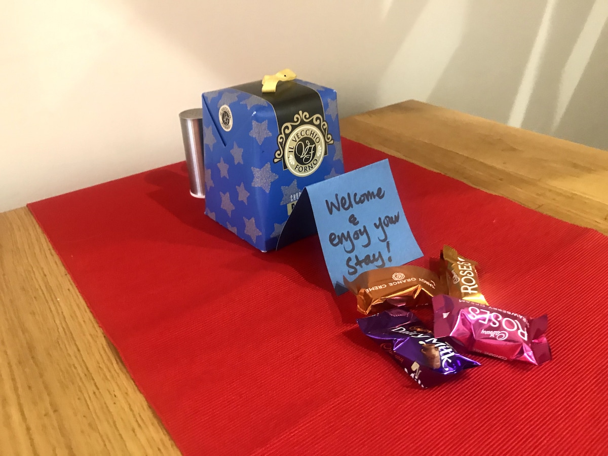 A small blue gift box, adorned with gold accents, rests on a red tablecloth. Next to the box, a colorful assortment of wrapped chocolates is displayed. A note, written on a blue sticky paper, offers a warm welcome to guests.