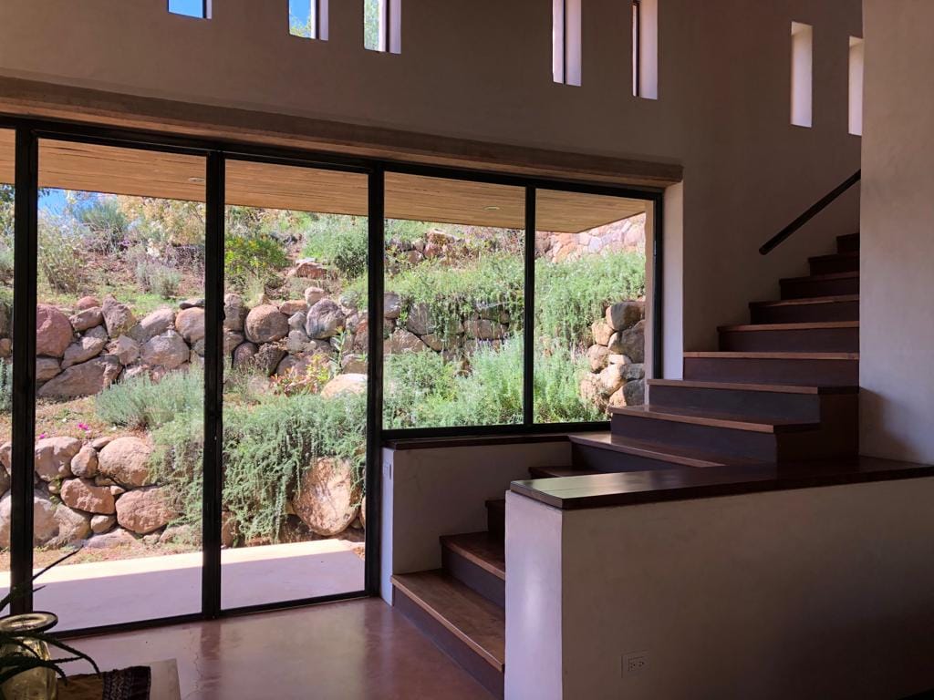 A modern interior space features large glass doors that frame views of a natural stone wall and vibrant greenery. A staircase with wooden steps leads to the upper level, enhancing the openness of the area. Natural light illuminates the interior, creating a bright atmosphere.