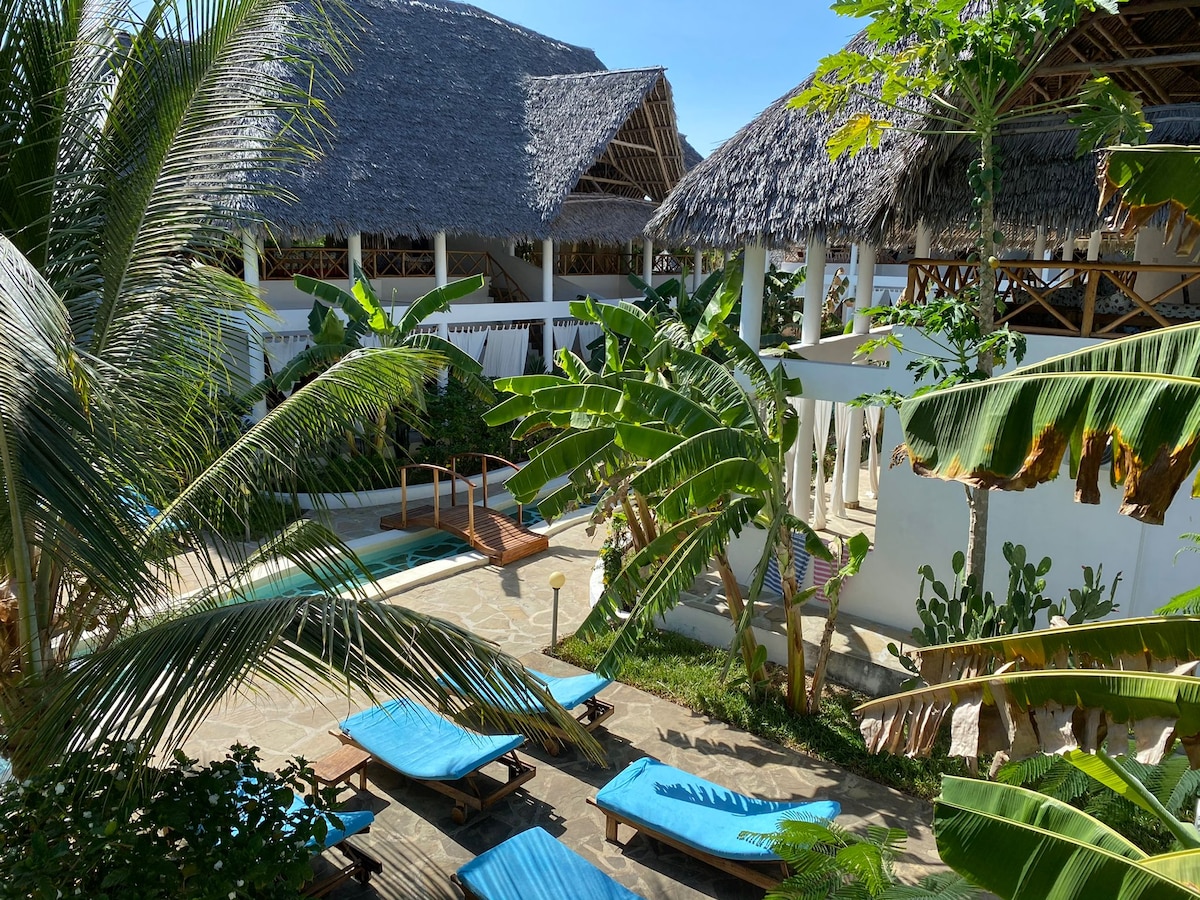 A tropical garden is showcased, featuring lush banana plants and palm trees surrounding a serene swimming pool. Sun loungers in a calming blue color are positioned for relaxation, creating a tranquil outdoor space. The architectural design includes thatched roofs, enhancing the natural aesthetic.