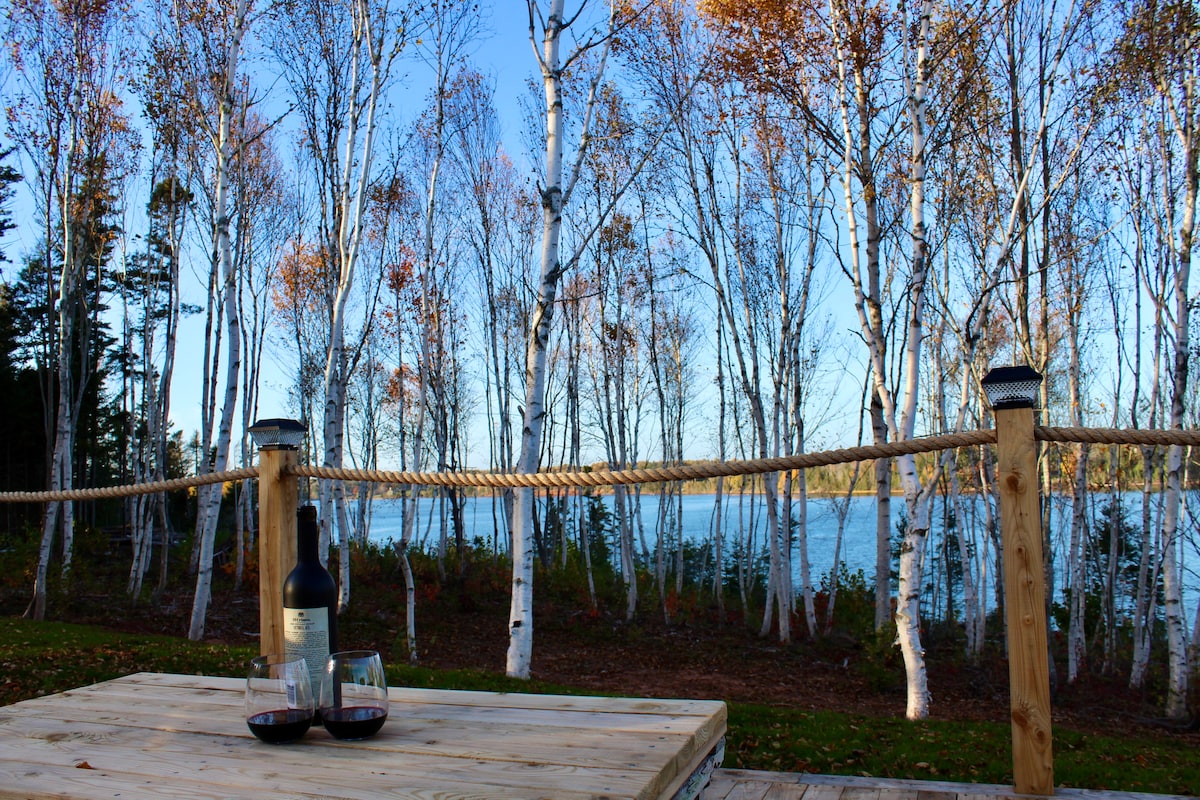 A wooden table is set with a bottle of wine and two glasses, positioned on a private deck overlooking a serene river. The background features a row of tall birch trees beside the water, with autumn foliage providing a colorful backdrop.