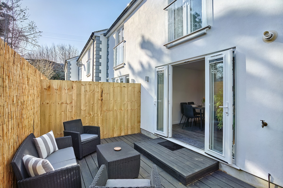 A private decking area is highlighted, featuring a comfortable seating arrangement with dark wicker furniture and soft cushions. The patio is enclosed by wooden fencing, providing a sense of seclusion. Double glass doors lead to the interior space, allowing natural light to fill the area.