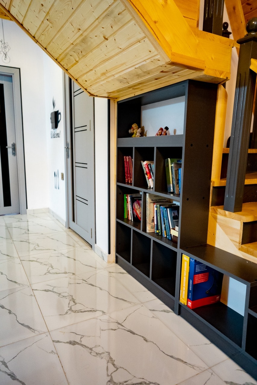 A sleek bookshelf is positioned against a wall, featuring various books and decorative items. The marble-patterned floor complements the modern design, while wooden stairs curve gracefully overhead. Neutral-colored doors are visible, adding to the contemporary ambiance of the entryway.