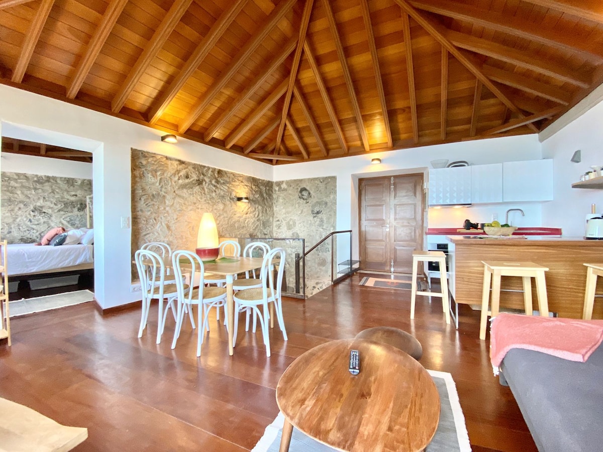 An open-plan living area features a wooden ceiling with exposed beams. A light wood dining table with four chairs sits adjacent to a modern kitchen. A cozy seating area includes two coffee tables and a sofa. Natural light fills the space from large windows.