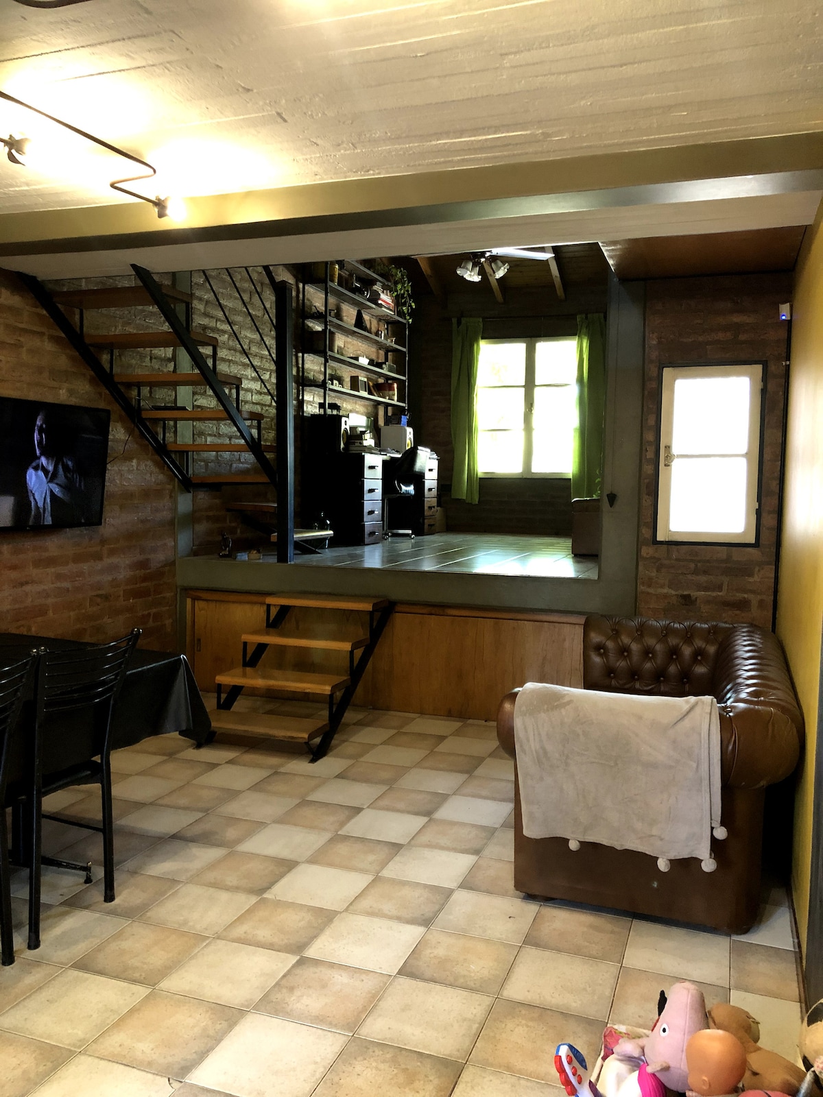 An open living area features a comfortable brown leather chair, and a dining set with black chairs. A staircase leads to a lofted space, while natural light enters through large windows adorned with light green curtains. A television screen is mounted on a wall.