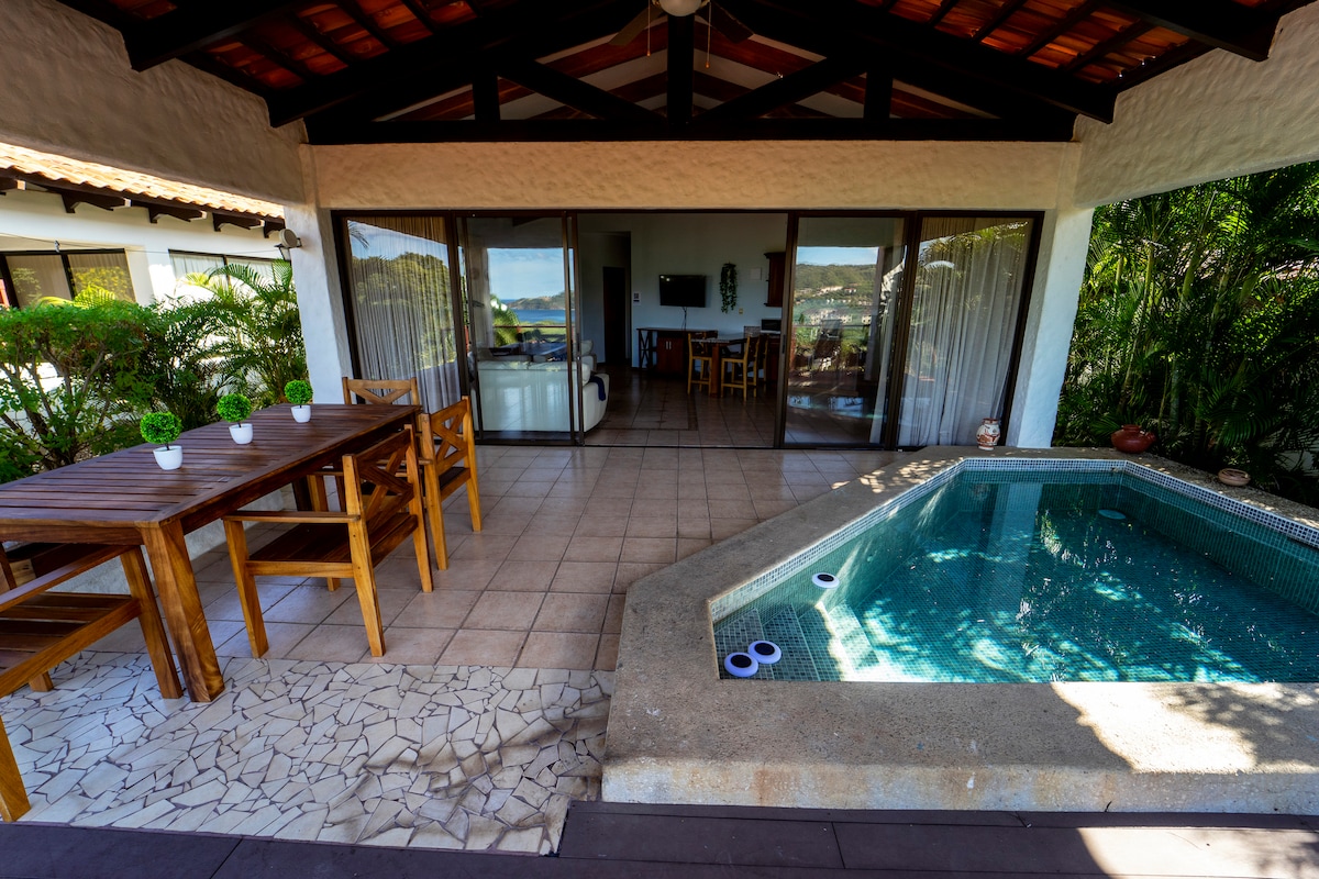 An outdoor area features a private plunge pool alongside a wooden dining table with matching chairs. Sliding glass doors offer a view of the interior space, allowing natural light to brighten the tiled floor, while surrounding greenery adds a touch of nature.