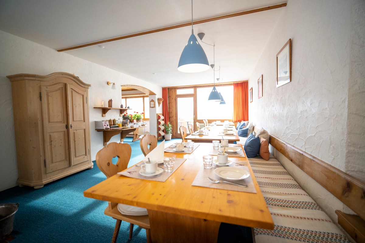 A dining area is presented with several wooden tables set for meals. Light blue pendant lights hang above, casting a soft glow. A cozy bench runs along the wall, adorned with cushions in varying tones. Large windows allow natural light to brighten the space.