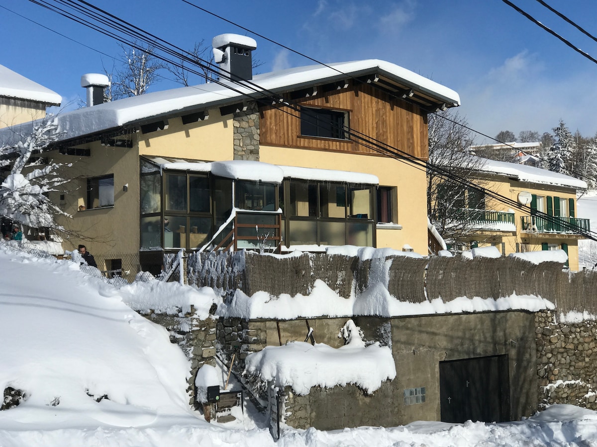 A modern structure with a combination of wooden and stone features is surrounded by snow. Large windows provide ample light inside, while a snow-covered roof adds to the winter atmosphere. Power lines are visible against a backdrop of the clear sky.