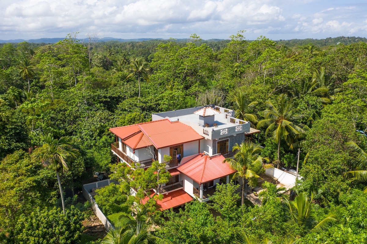 An aerial view captures the Tree Trail Villa surrounded by lush greenery, with a striking contrast between the red roofs and the vibrant jungle. The structure showcases multiple levels, with spacious balconies offering views of the natural landscape.