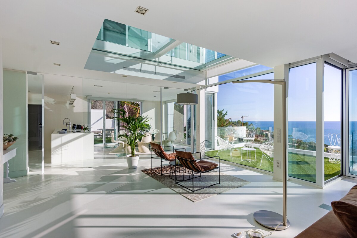 An airy living area is illuminated by natural light, featuring large glass windows that offer expansive ocean views. Comfortable seating is arranged on a neutral-toned rug, alongside a stylish lamp. The open layout connects to a patio showcasing greenery and outdoor furnishings.