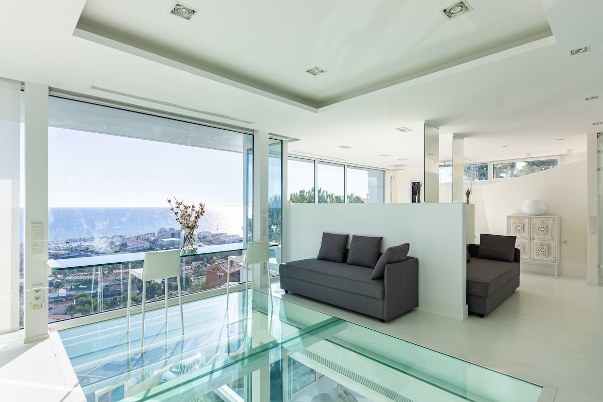 A spacious living area features floor-to-ceiling glass walls that offer expansive views of the sea. Light-colored furnishings, including a dark gray sofa, complement the bright interior design. A glass dining table is positioned near the windows, enhancing the open feel of the space.