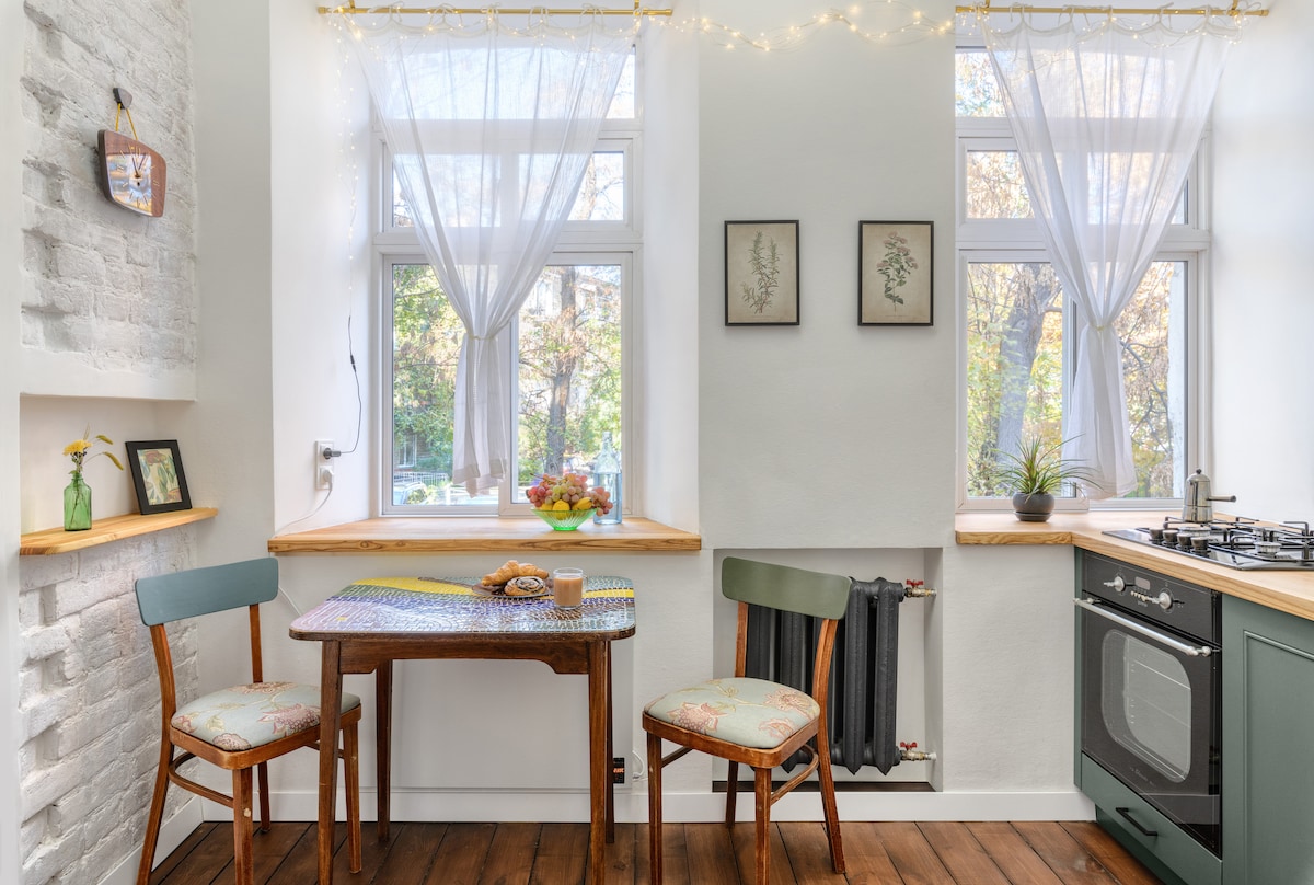 A well-lit kitchen area features two wooden chairs around a table, with light-colored curtains framing the windows. Fresh flowers are placed on the windowsill, and a gas stove is positioned next to the colorful cabinetry. A rustic charm complements the modern finishes.