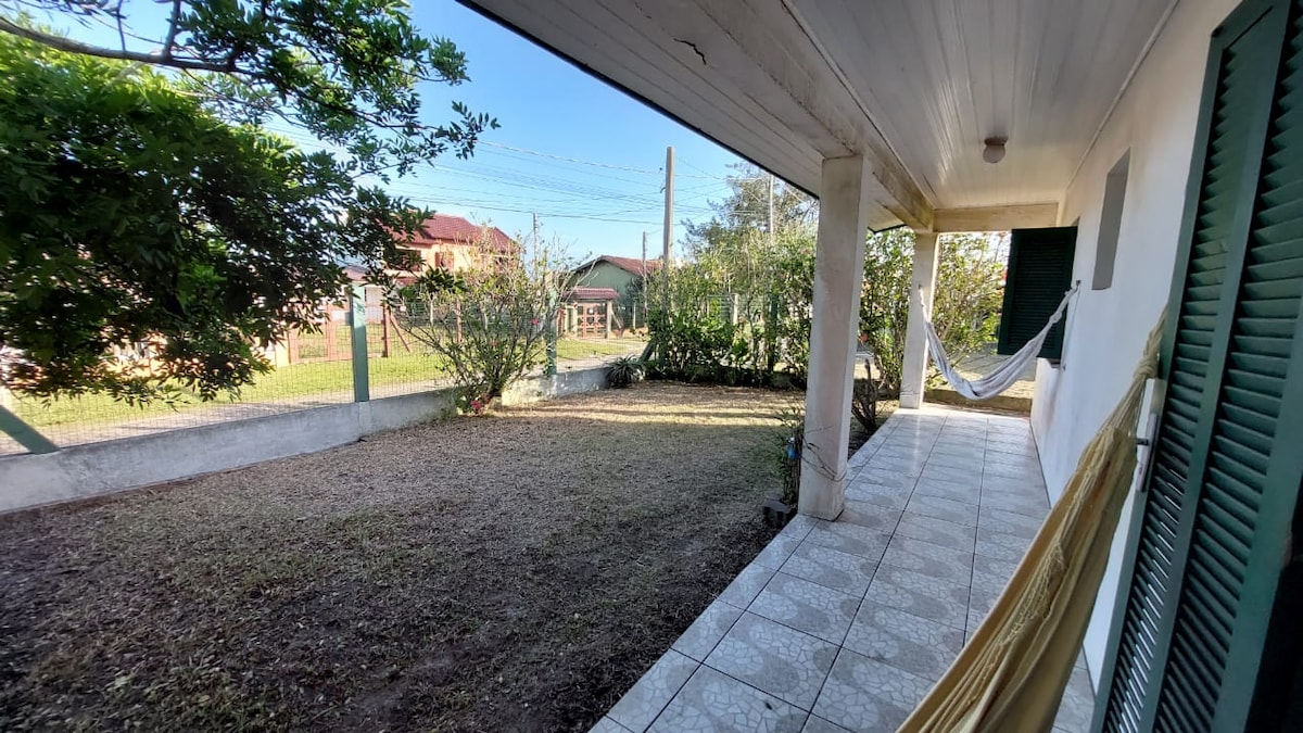 A spacious porch is decorated with a relaxed hammock, providing a comfortable lounging area. Green shrubs and small trees border the yard, enhancing the outdoor space. A tiled pathway leads to the entrance, with natural light illuminating the scene.