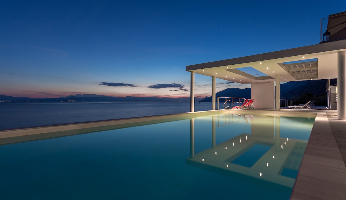 An infinity pool stretches toward the horizon, reflecting the evening sky. Soft lighting illuminates the surrounding area, enhancing the tranquil atmosphere. Comfortable lounge chairs are positioned nearby, inviting relaxation and appreciation of the scenic coastal view.