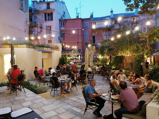 An outdoor dining area is depicted with scattered tables and chairs under string lights, creating a cozy ambiance. Patrons are seen enjoying their meals, surrounded by lush greenery and softly illuminated walls of adjacent buildings. The scene evokes a relaxed evening atmosphere.