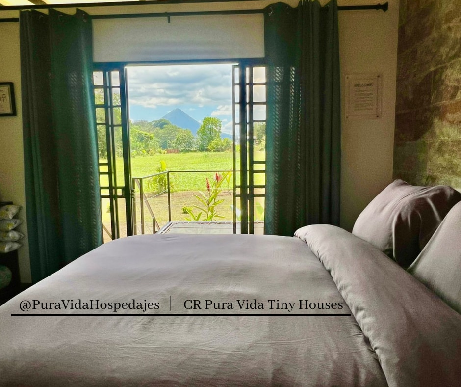 A spacious bed with soft linens is positioned near large open doors, offering a clear view of lush greenery and the Arenal Volcano in the distance. Natural light fills the room, creating a relaxing atmosphere framed by patterned curtains.