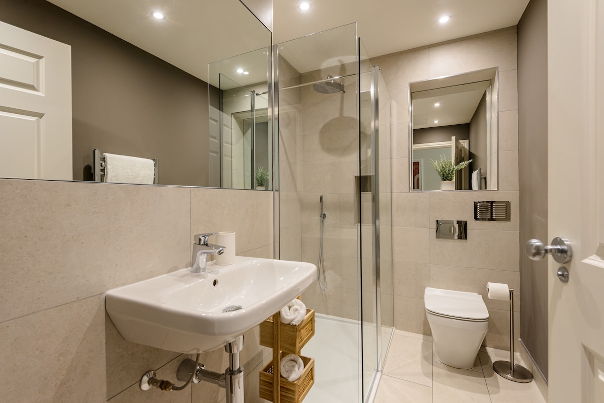 The modern bathroom features a walk-in shower with clear glass doors, a sleek sink, and a toilet. Neutral tiles adorn the walls and floor, complemented by soft ambient lighting. A decorative plant sits atop a recessed shelf, adding a touch of greenery to the space.