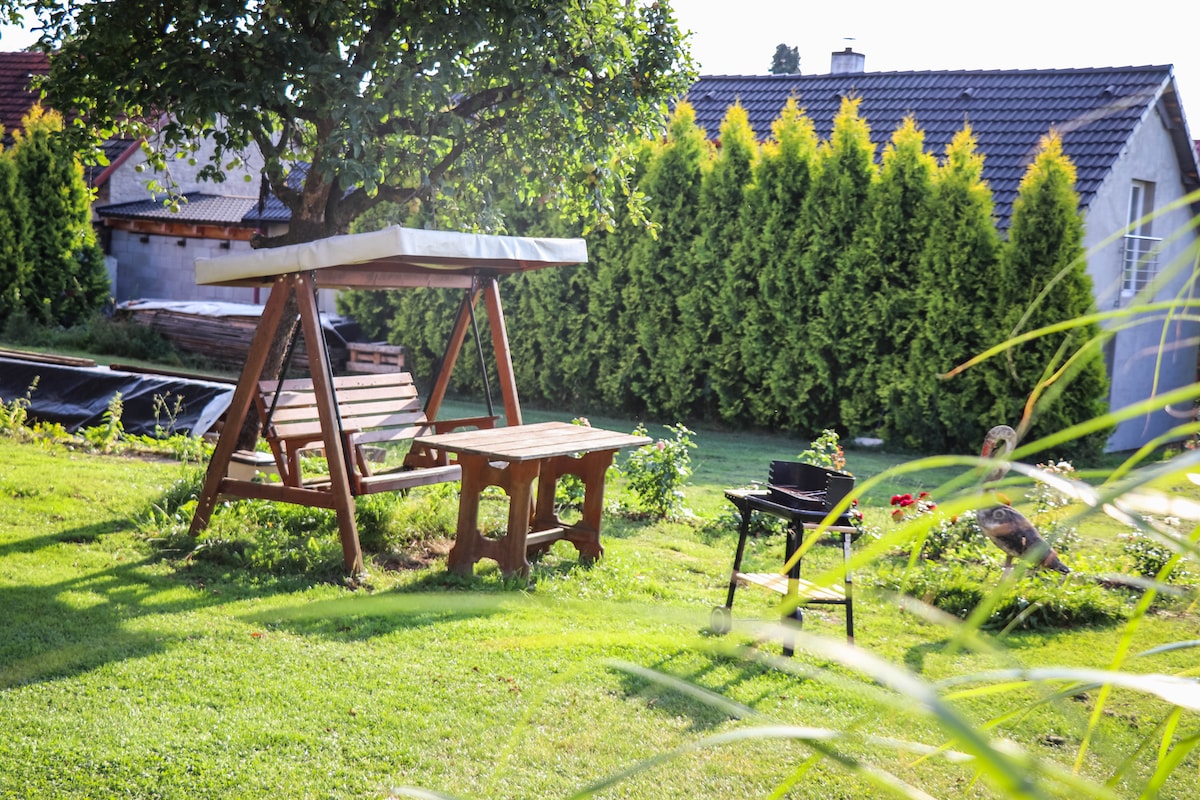 A wooden swing sits beneath a tree, accompanied by a small table nearby. Lush greenery surrounds the space, creating a tranquil atmosphere. A grill is positioned on the lawn, ready for outdoor cooking. The landscape reflects a well-maintained garden environment.