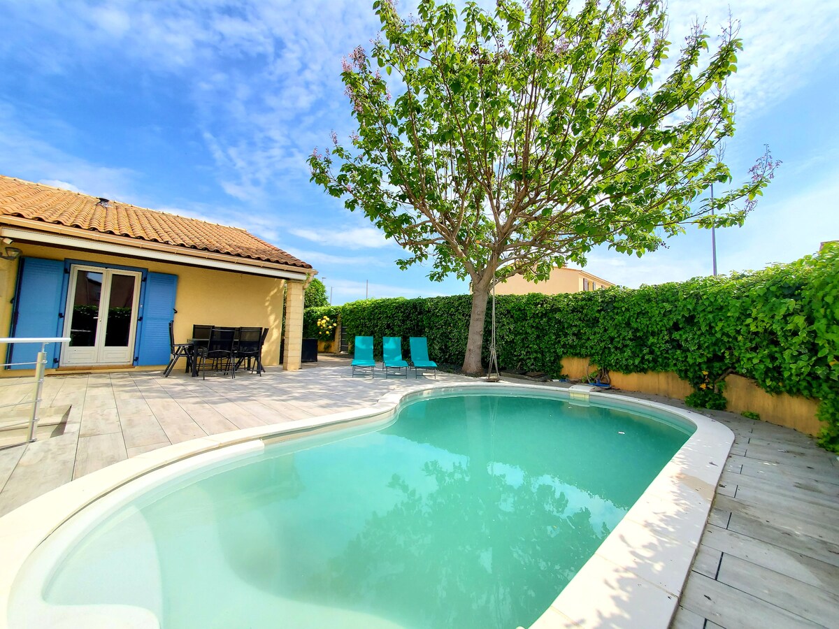 A serene outdoor space features a private pool surrounded by a wooden deck. Comfortable seating options are available, including green lounge chairs. Lush greenery provides privacy, while a shaded tree adds a natural element to the setting. The exterior of the villa is visible, showcasing bright blue shutters.