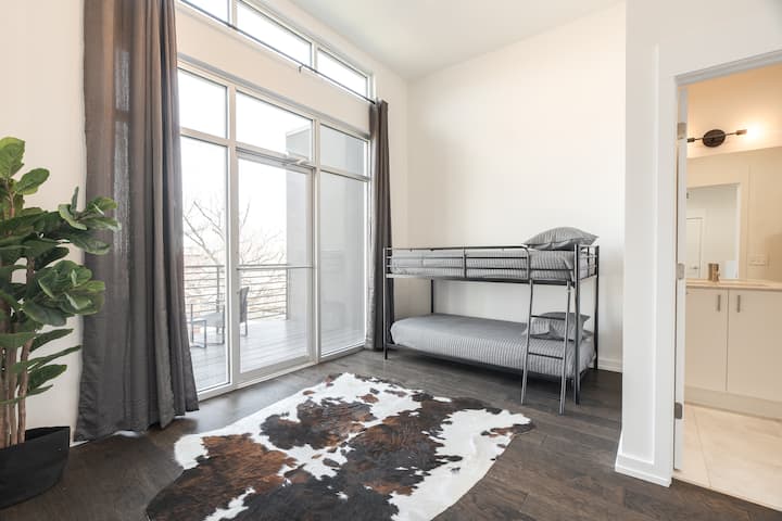 Minimalist bedroom with metal bunk bed, cowhide rug, and dark wood floors. Floor-to-ceiling windows open to a balcony, flooding the space with light. A potted plant and high ceiling add airy, modern charm.