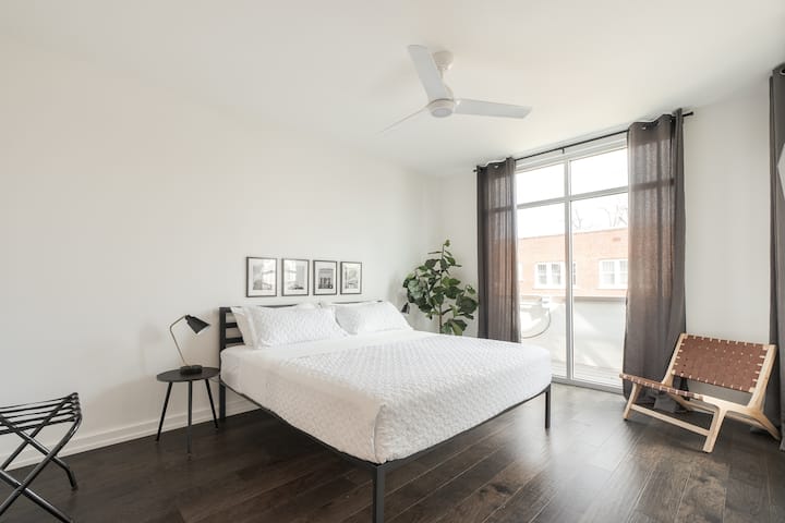 Minimalist bedroom with white bedding, black metal frame, and geometric wall art. Dark wood floors, sheer curtains, and natural light create a serene, modern escape with elegant simplicity.