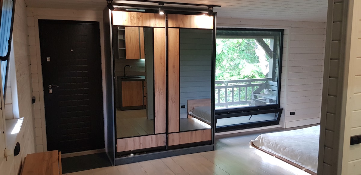 The interior of a cozy studio features wooden accents and large mirrored sliding wardrobes. A window offers a view of surrounding greenery, while a light-colored mattress is placed on a wooden platform. The kitchenette is subtly visible in the background.
