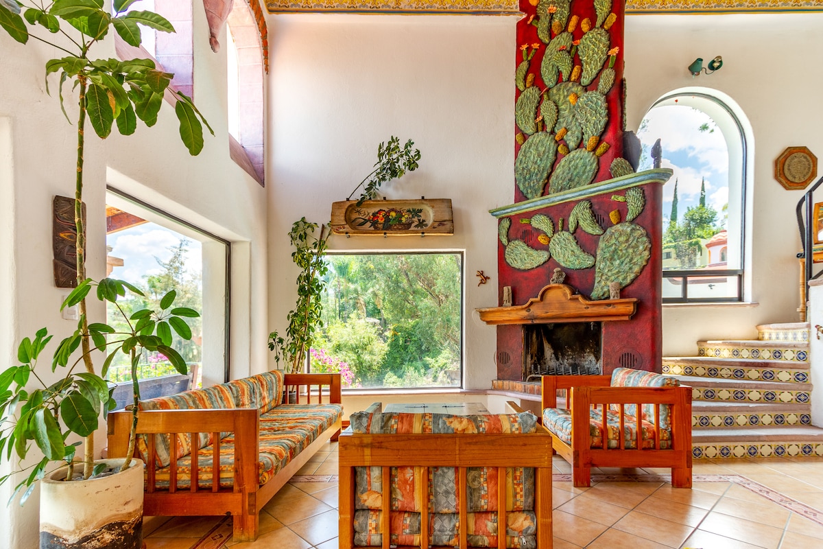 A cozy seating area is highlighted by colorful patterned sofas and a wooden coffee table. A fireplace sits beneath a decorative wall featuring cacti artwork. Large windows allow natural light to illuminate the space, creating a connection with the greenery outside.