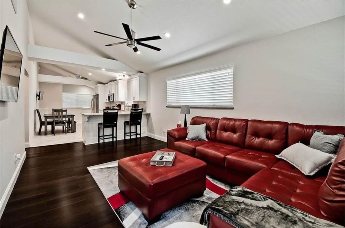 A modern living space features a large red sectional sofa and a matching ottoman, positioned on a patterned rug. A sleek dining area can be seen in the background, with a table and chairs. Natural light enters through a window adorned with blinds.