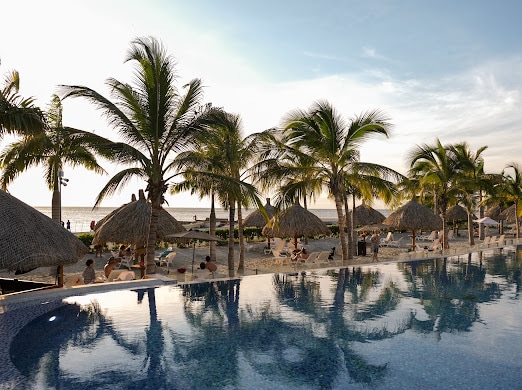 A serene beach setting is captured with palm trees surrounding a tranquil swimming pool. Thatched umbrellas are positioned on the sand, where guests can be seen relaxing. The ocean horizon meets the sky at sunset, creating a calming atmosphere.