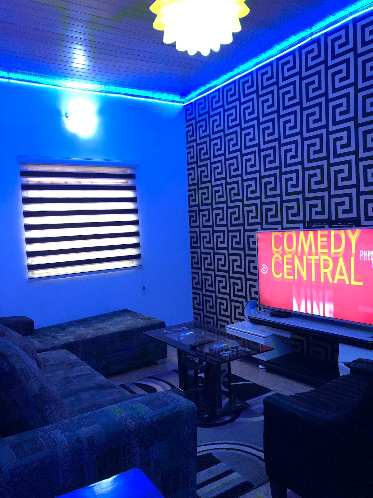 A cozy living area features a dark patterned accent wall alongside vibrant blue ambient lighting. A flat-screen television is situated on a media unit, displaying a channel name. Comfortable seating options, including a sofa and chairs, are arranged around a glass coffee table.