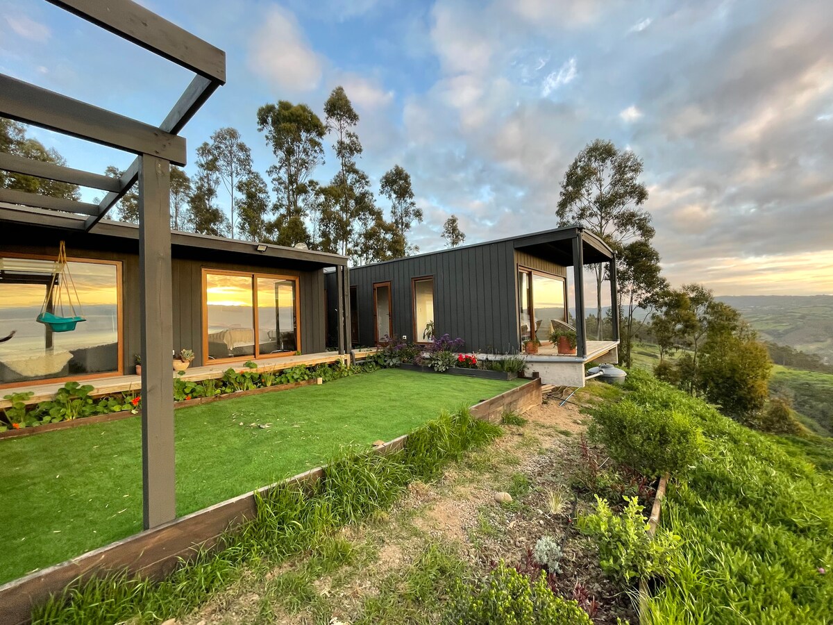 A modern house is situated on a hillside, surrounded by lush greenery and flowering plants. Large windows provide views of the landscape, while a small terrace extends from the structure. The sky reflects soft colors from the setting sun, enhancing the tranquil setting.
