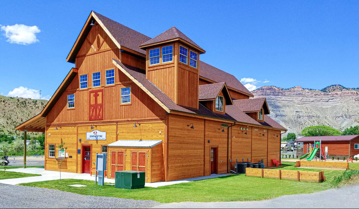 A spacious wooden building features a prominent gabled roof and multiple windows allowing natural light. Nearby, a playground is visible, along with grassy areas. The structure is set against a backdrop of hills, emphasizing its rustic charm.