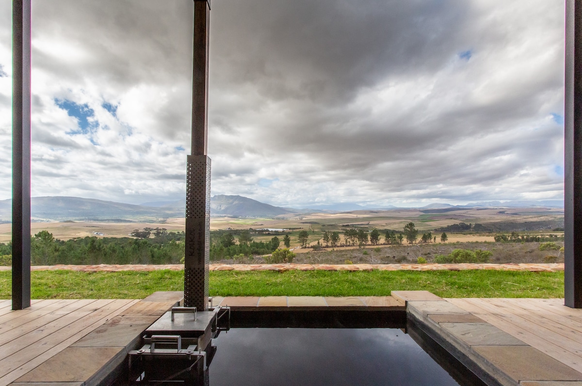 A wood-fired hot tub is positioned on a wooden deck, overlooking expansive views of the rolling hills and valleys below. The cloudy sky adds depth to the landscape, while greenery borders the foreground, creating a serene environment.