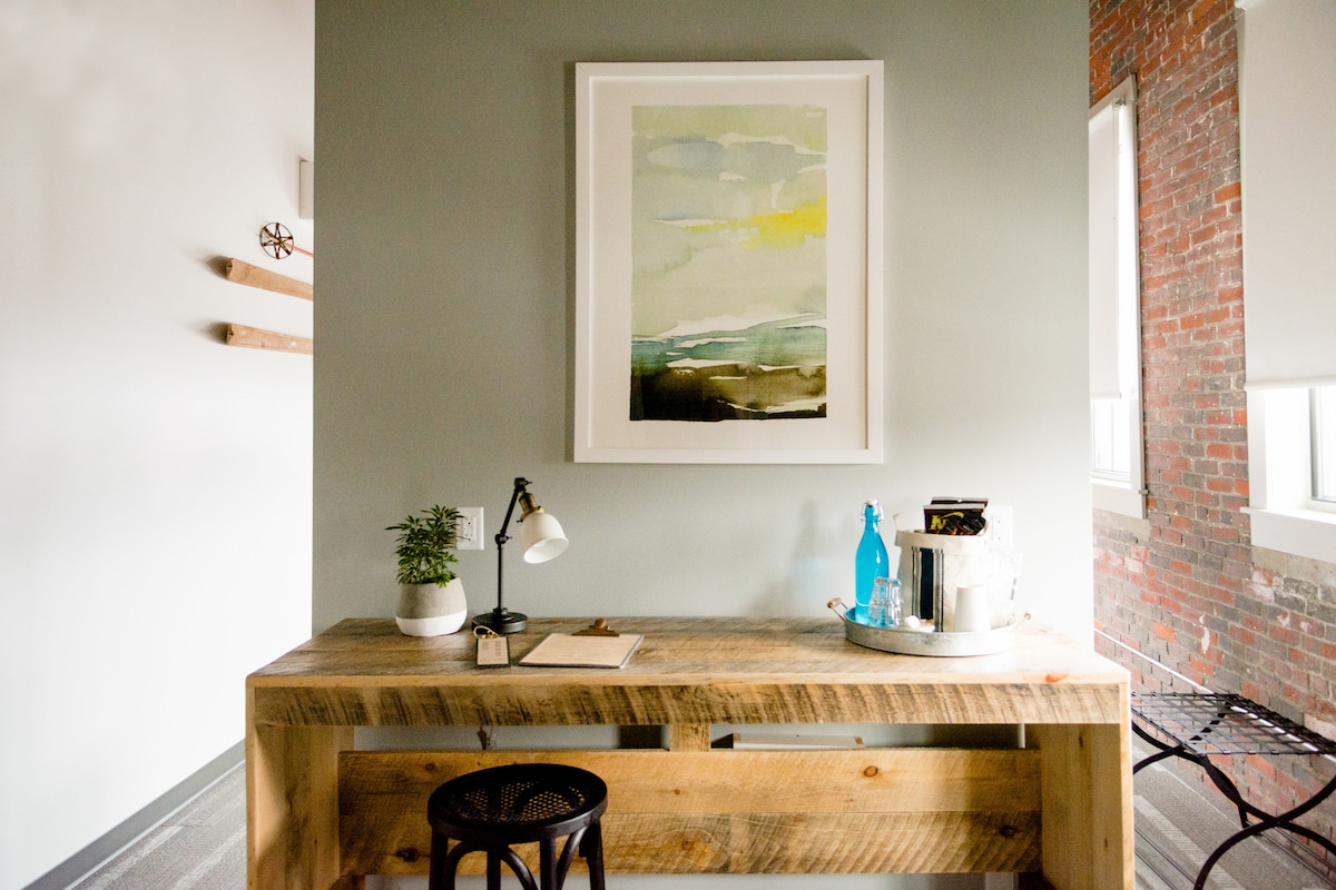 A custom reclaimed wood desk is paired with a circular black stool, positioned under a wall-mounted artwork. A small plant, a notepad, a lamp, and a water bottle are placed on the desk. Exposed brick can be seen in the background, enhancing the room's character.