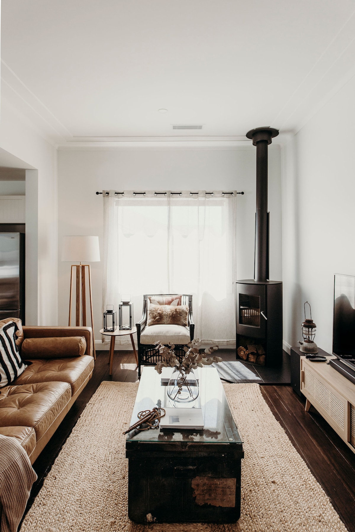 A cozy living area is highlighted by a warm wood-burning stove and a large glass coffee table. The earthy tones of the leather sofa complement the soft texture of the area rug. Natural light enters through sheer curtains, creating an inviting space.