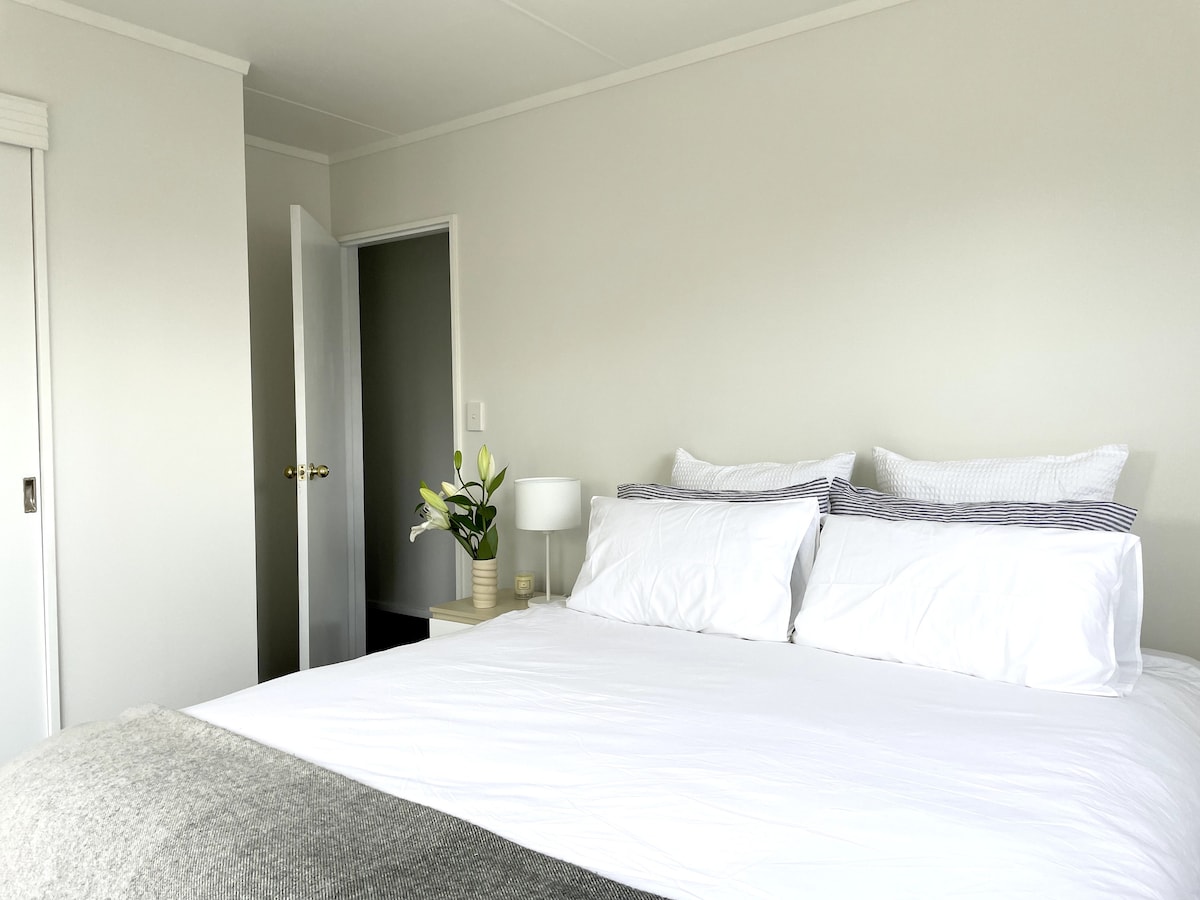 A bright and airy bedroom features a large bed with crisp white linens and accented pillows. A bedside table holds a lamp and a vase with fresh flowers. A door leads to another room, enhancing the light and open feel.