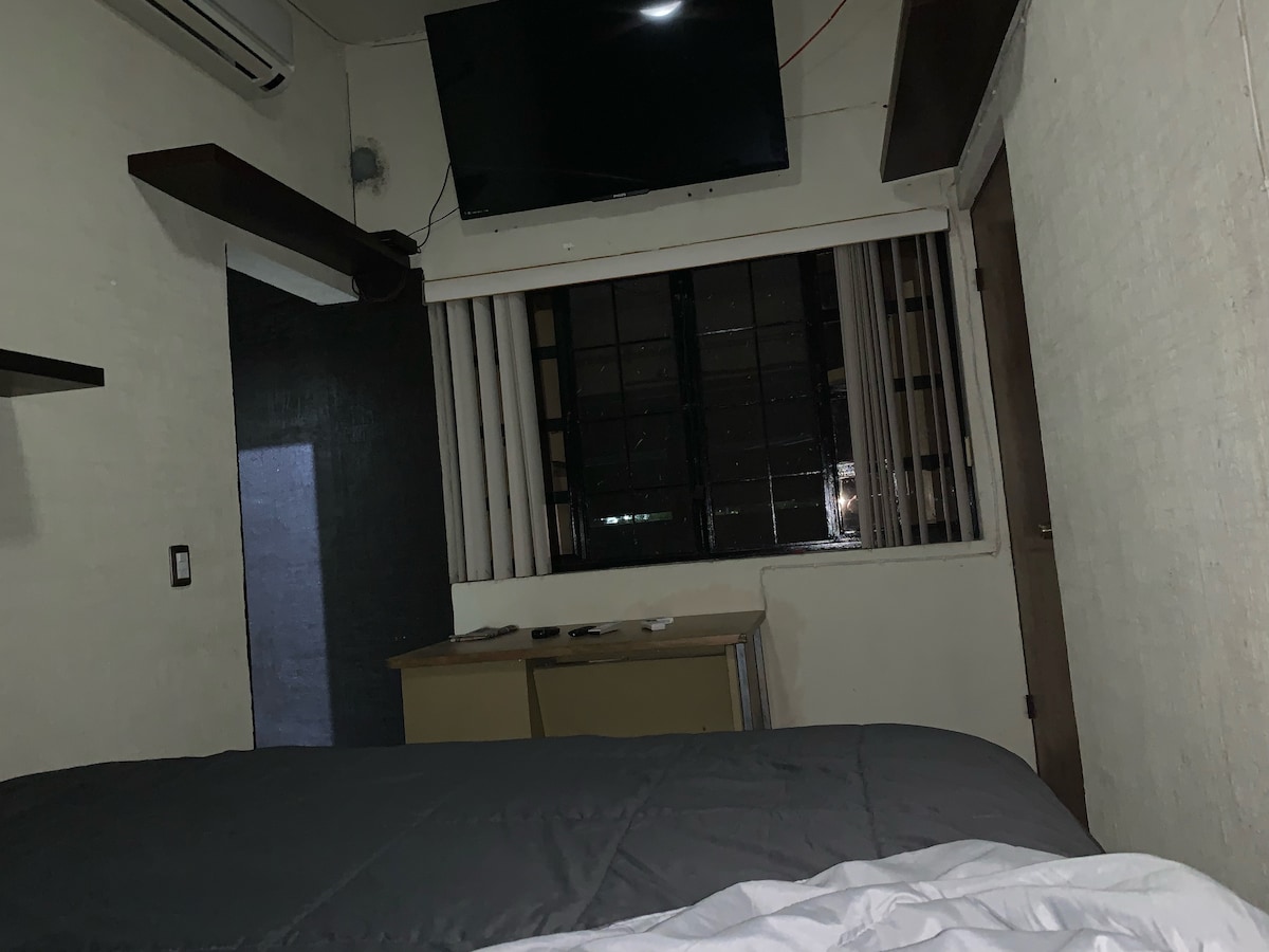 A sleeping area is presented with a spacious bed draped in a gray comforter. A flat-screen TV is mounted on the wall opposite a window with vertical blinds. A wooden desk with papers is positioned nearby, contributing to the functional layout of the room.