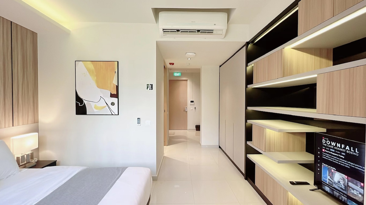 A modern bedroom area features a spacious bed dressed in soft linens, complemented by bedside lighting. A flat-screen TV is positioned on a sleek, minimalist shelf, while a framed artwork adds a touch of style. The soft neutral tones create a calming environment.