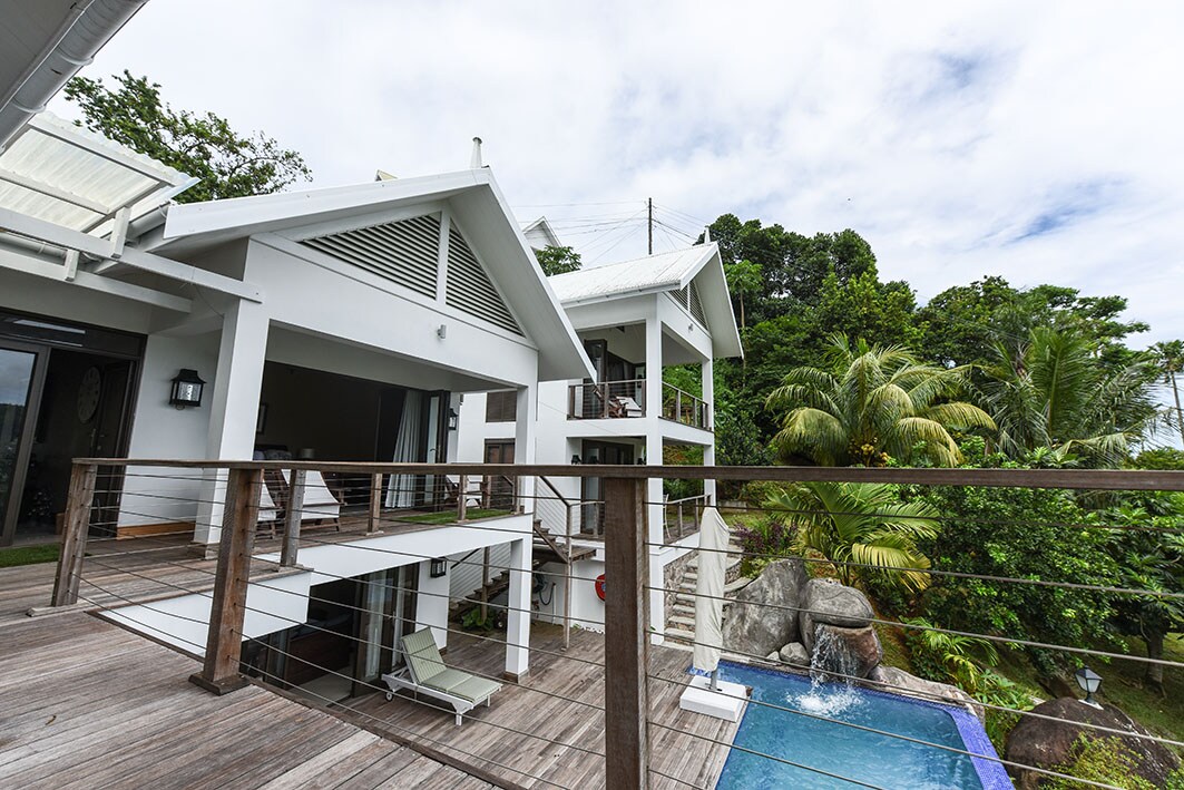 An upper-level balcony extends from the apartment, featuring wooden flooring and a railing. Views of lush greenery and a small pool with a water feature are visible. The structure exhibits a modern design with sloped roofs and large windows, inviting natural light into the space.