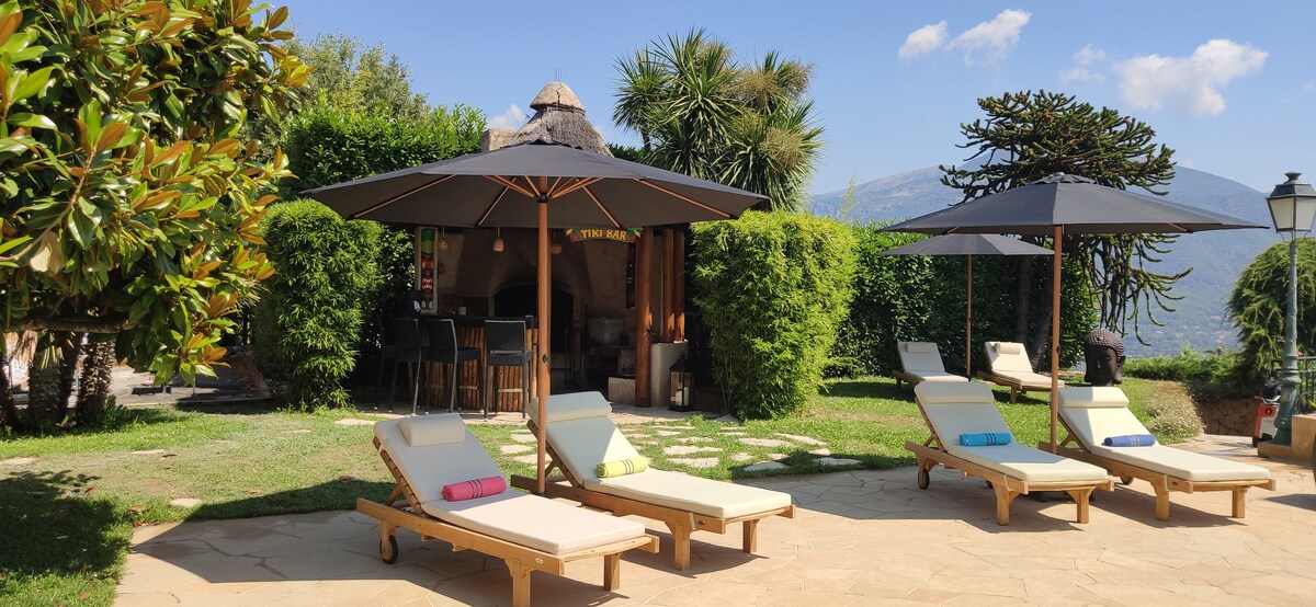 A spacious outdoor area features multiple sun loungers arranged on a patterned stone surface. Large umbrellas provide shade, while tropical plants and trees surround the space. In the background, a shaded dining area is visible, creating a relaxing environment for guests.