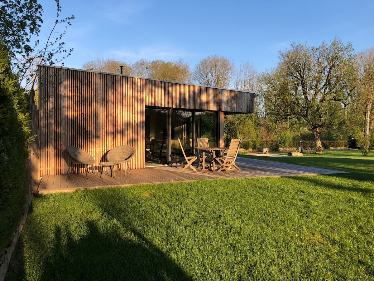 A modern wooden structure is surrounded by lush green grass. Two seating areas are present on the deck, facing a tranquil outdoor space. Large glass doors grant views of the interior, while trees and foliage create a serene backdrop.