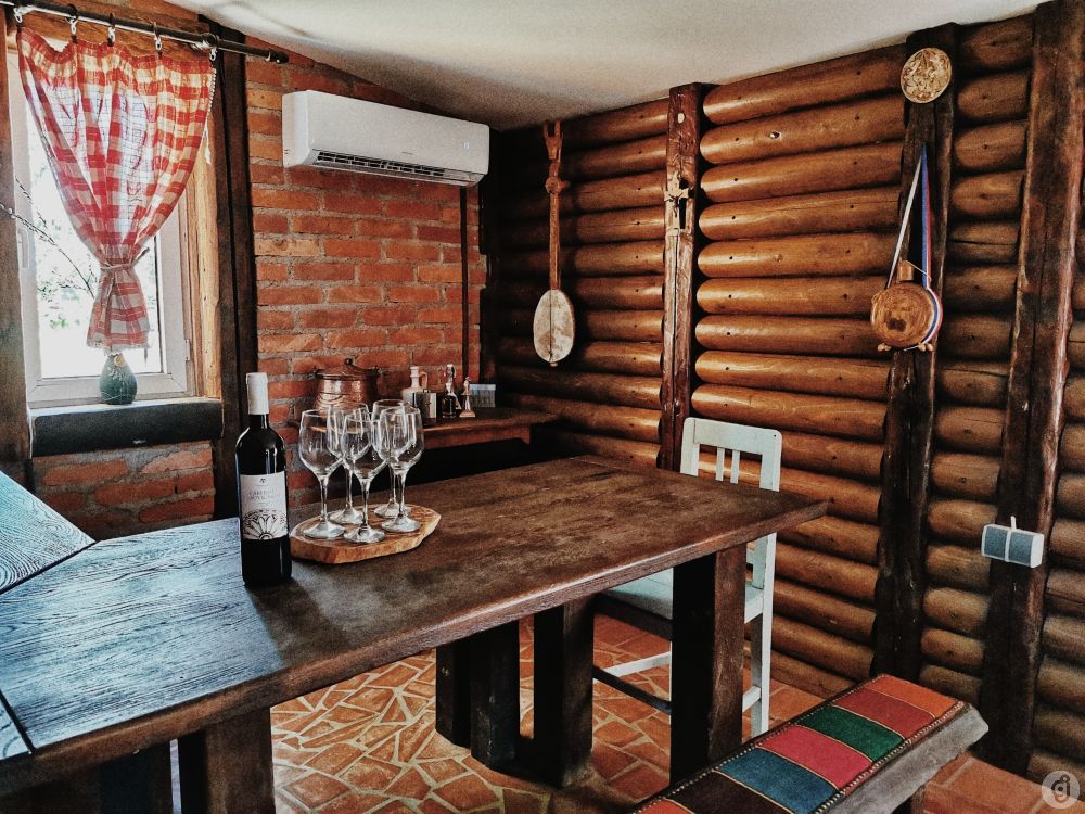 An interior dining space features a wooden table surrounded by light-colored chairs. Wine glasses are neatly arranged on a wooden tray beside a bottle of wine. The walls are adorned with rustic wooden logs, and a window is dressed with red checkered curtains.