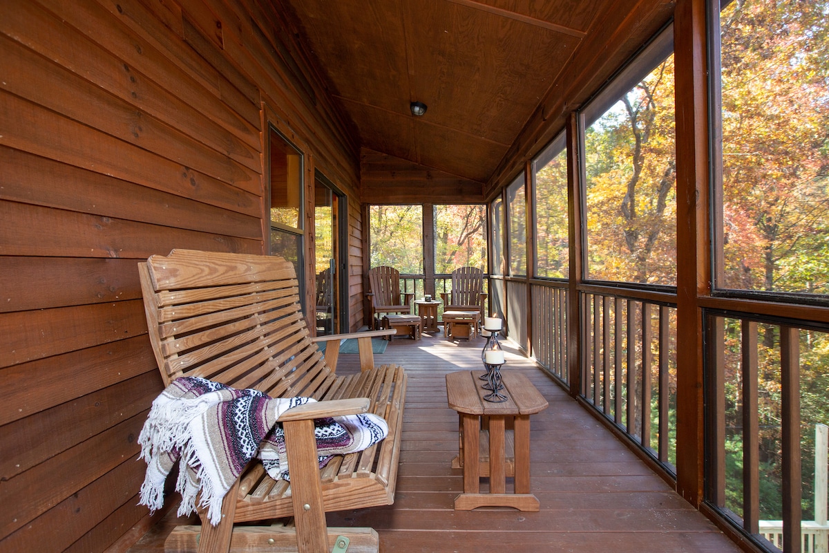 A spacious screened-in porch is visible, featuring wooden rocking chairs and a small table. The warm wood tones of the cabin complement the natural surroundings, with trees displaying autumn colors in the background. Soft sunlight filters through the screens, creating a cozy atmosphere.