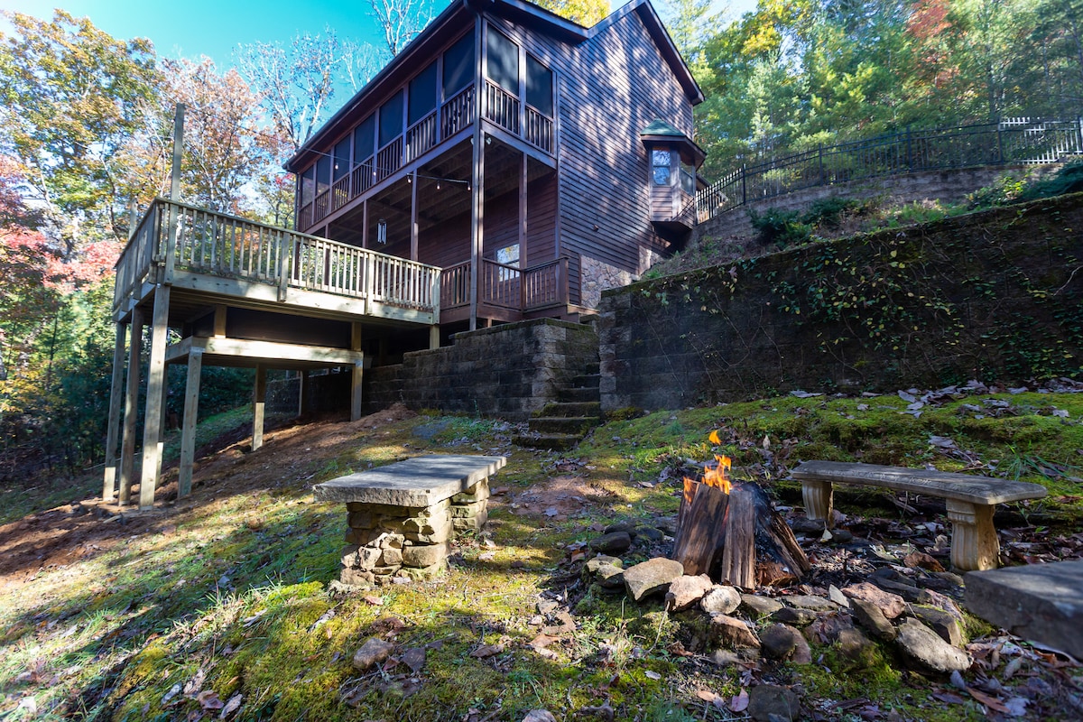 The chalet's exterior is shown, set against a backdrop of trees. A spacious deck with several entry points is visible above stone steps. A fire pit with stone seating is located in the foreground on the lush, green ground, enhancing the outdoor ambiance.